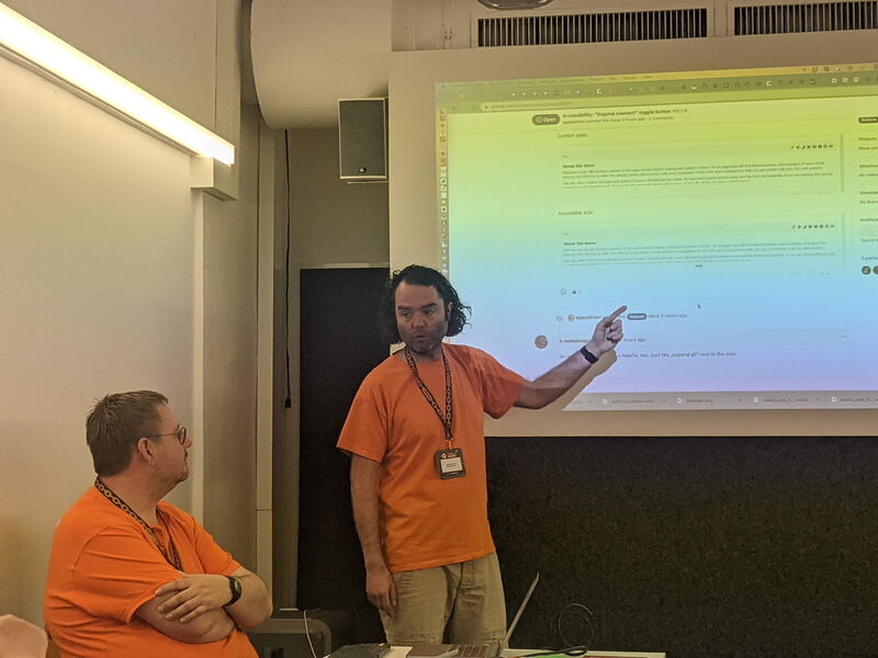 Zwei Männer in orangen Contao-Shirts vor einer projizierten GitHub-Seite, einer zeigt auf ein Issue zur Barrierefreiheit