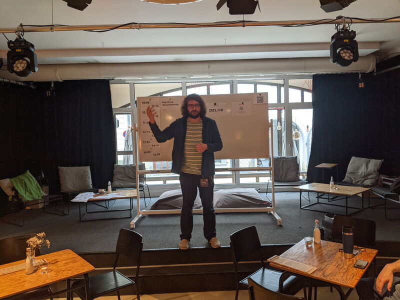 Ein Mann steht auf der Bühne des Hauptraums vor dem Whiteboard mit dem Zeitplan, im Raum Sessel, Sitzkissen und Scheinwerfer