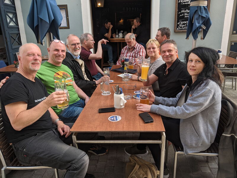 Neun Leute sitzen um einen langen Tisch im Freien vor einem Wirtshaus, Gläser in der Hand, Blick in die Kamera