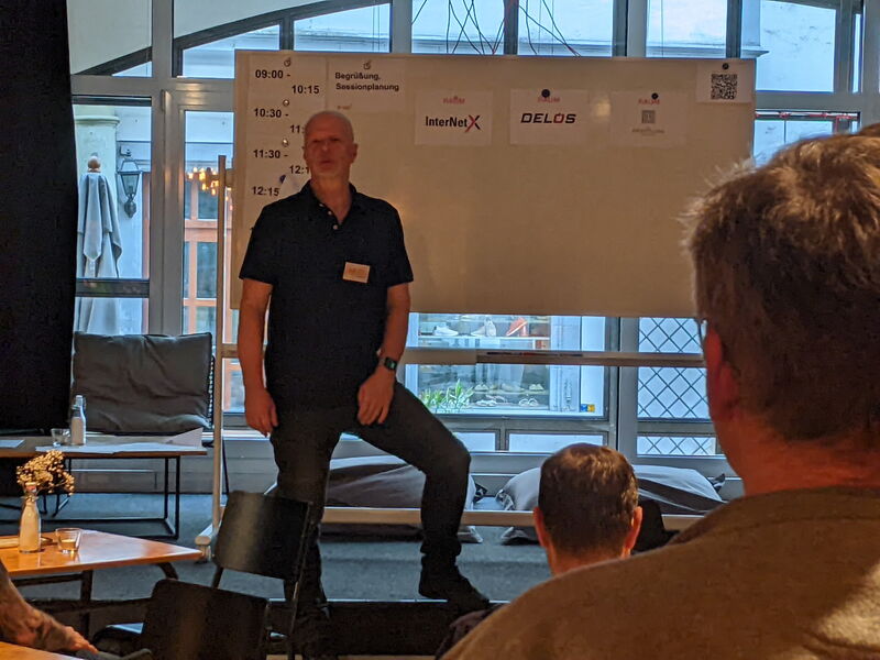 Ein Mann mit Namensschild steht vor dem Whiteboard mit dem Tagesplan, davor Zuhörer