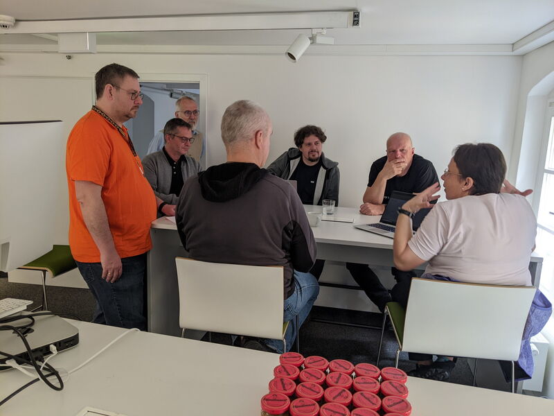Sechs Leute stehen und sitzen um einen Tisch mit Laptop, im Vordergrund die Senfgläser
